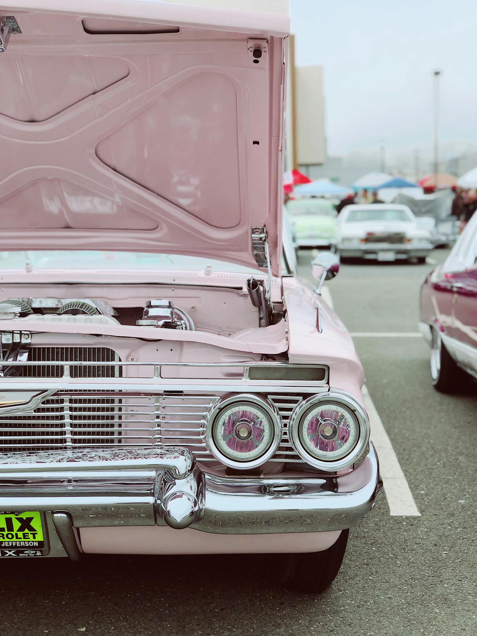 Front view of a pink vintage car with an open hood at an outdoor auto show.
